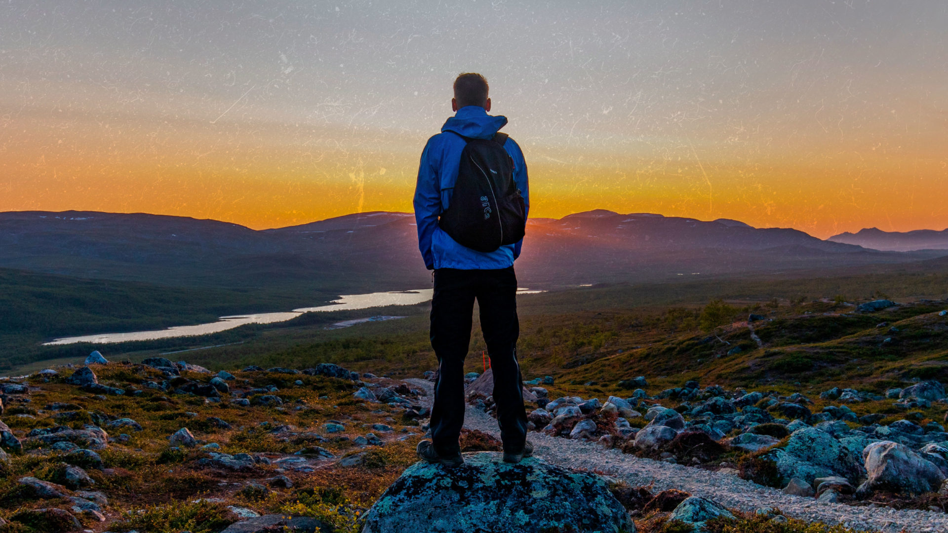 En filmaffisch där en man klädd i blåa vandringskläder står med ryggen mot kameran. Han står på en sten och betraktar solnedgången över fjällen.