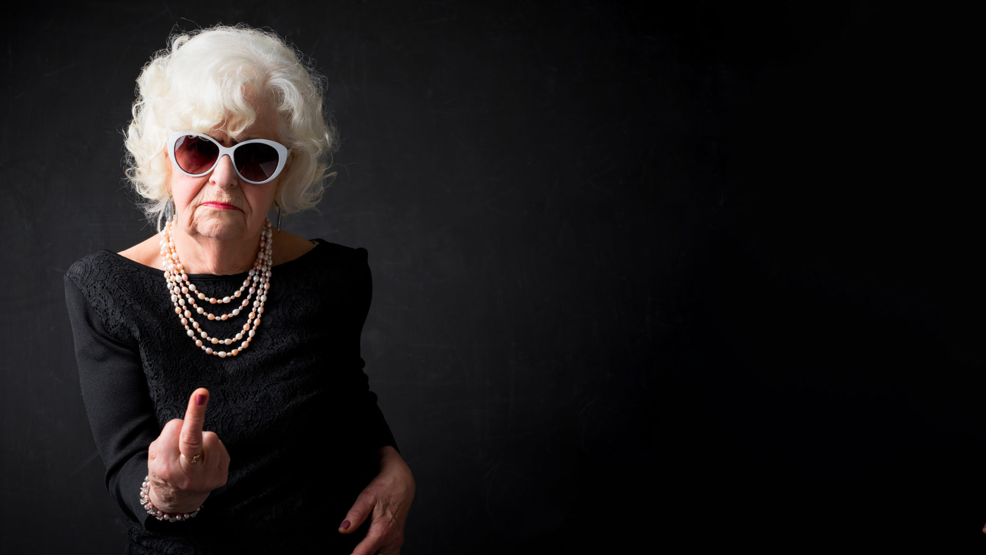 A film poster depicting an elderly woman who is facing the camera and giving the finger. She has short gray hair and she is wearing a black dress, sunglasses and a pearl necklace.