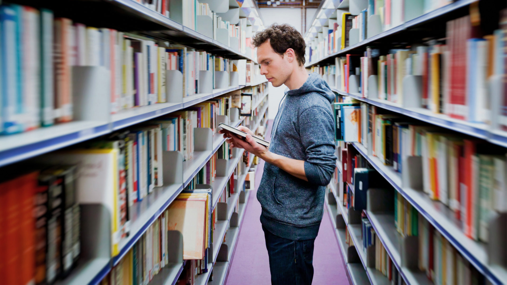 A man in his twenties, wearing a sweatshirt, is standing between two library shelves. He is reading a book.