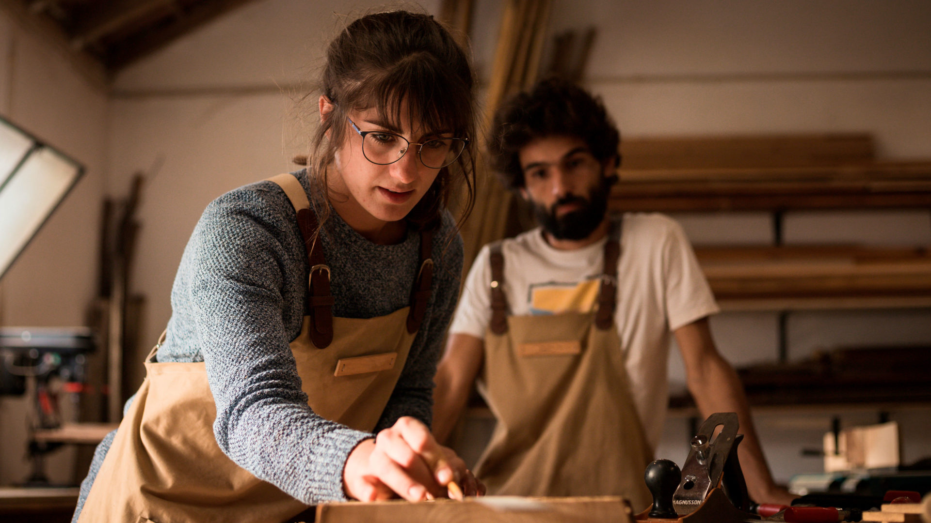 En ung brunhårig kvinna med glasögon synar ett hantverk av trä. En ung man med skägg betraktar henne uppmärksamt i bakgrunden.