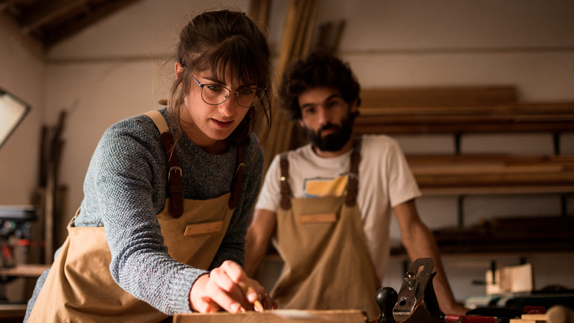 A young brown-haired woman with glasses is examining a piece of wood in a woodworking class. In the background, a young bearded man is looking at her.
